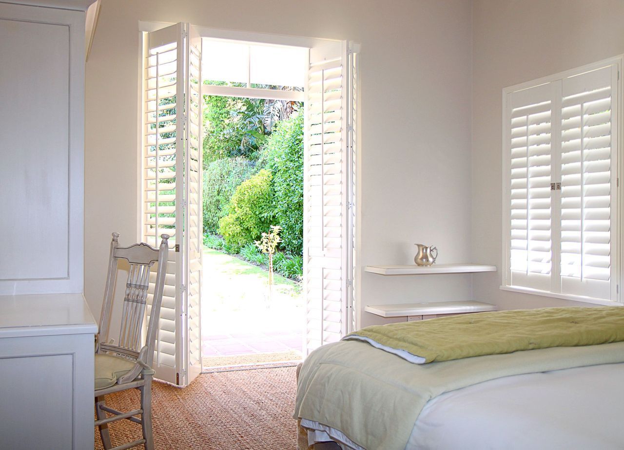A bright bedroom with white walls, a bed with a sage green quilt, a white chair, and doors opening to a garden.