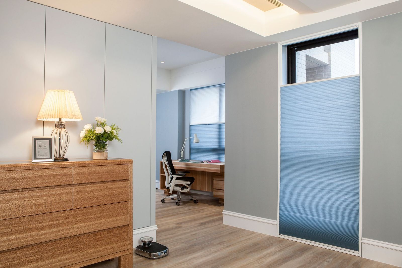 A modern room with a wood dresser, table lamp, and blue pleated blinds on a tall window, with an office visible beyond.