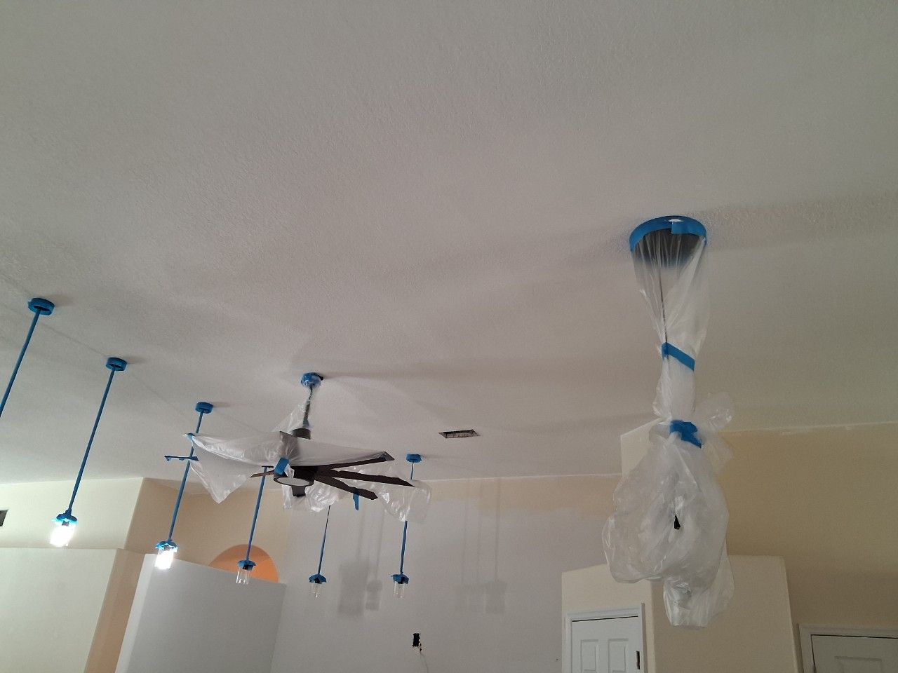 A ceiling being painted white, with light fixtures and a ceiling fan covered in plastic and blue painter's tape.