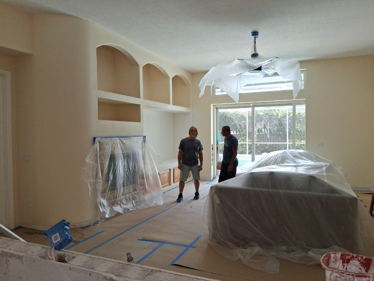Two people stand in a living room undergoing renovation, with furniture covered in plastic and blue painter's tape on floors.