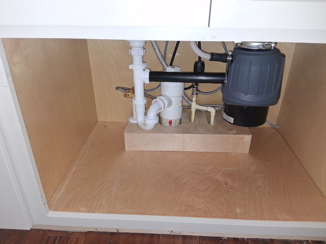 Under-sink kitchen cabinet showing a garbage disposal unit, plumbing pipes, and a small wooden riser block.