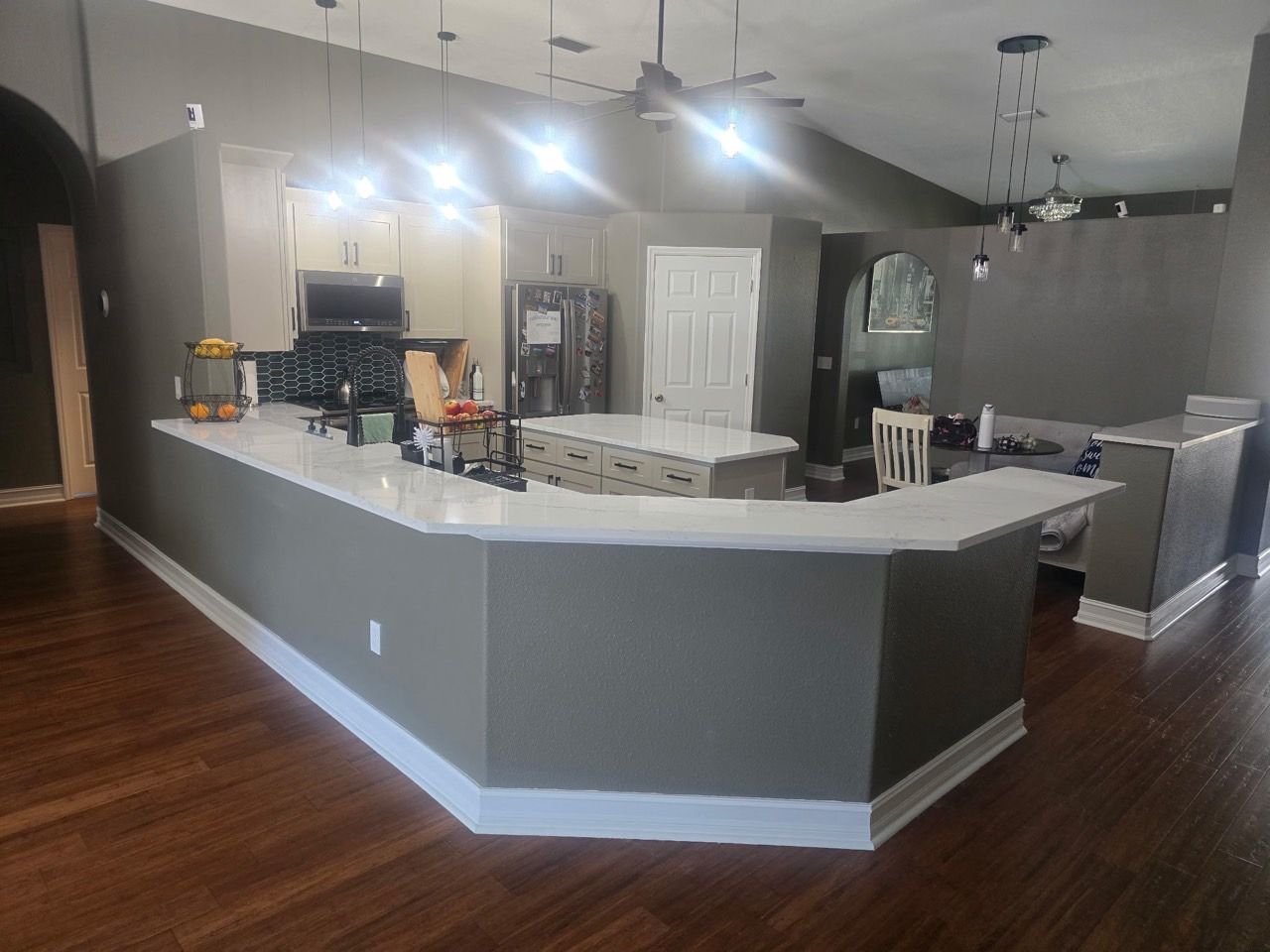 A modern kitchen with gray walls, white counters, wood floors, and hanging pendant lights, viewed from the living area.
