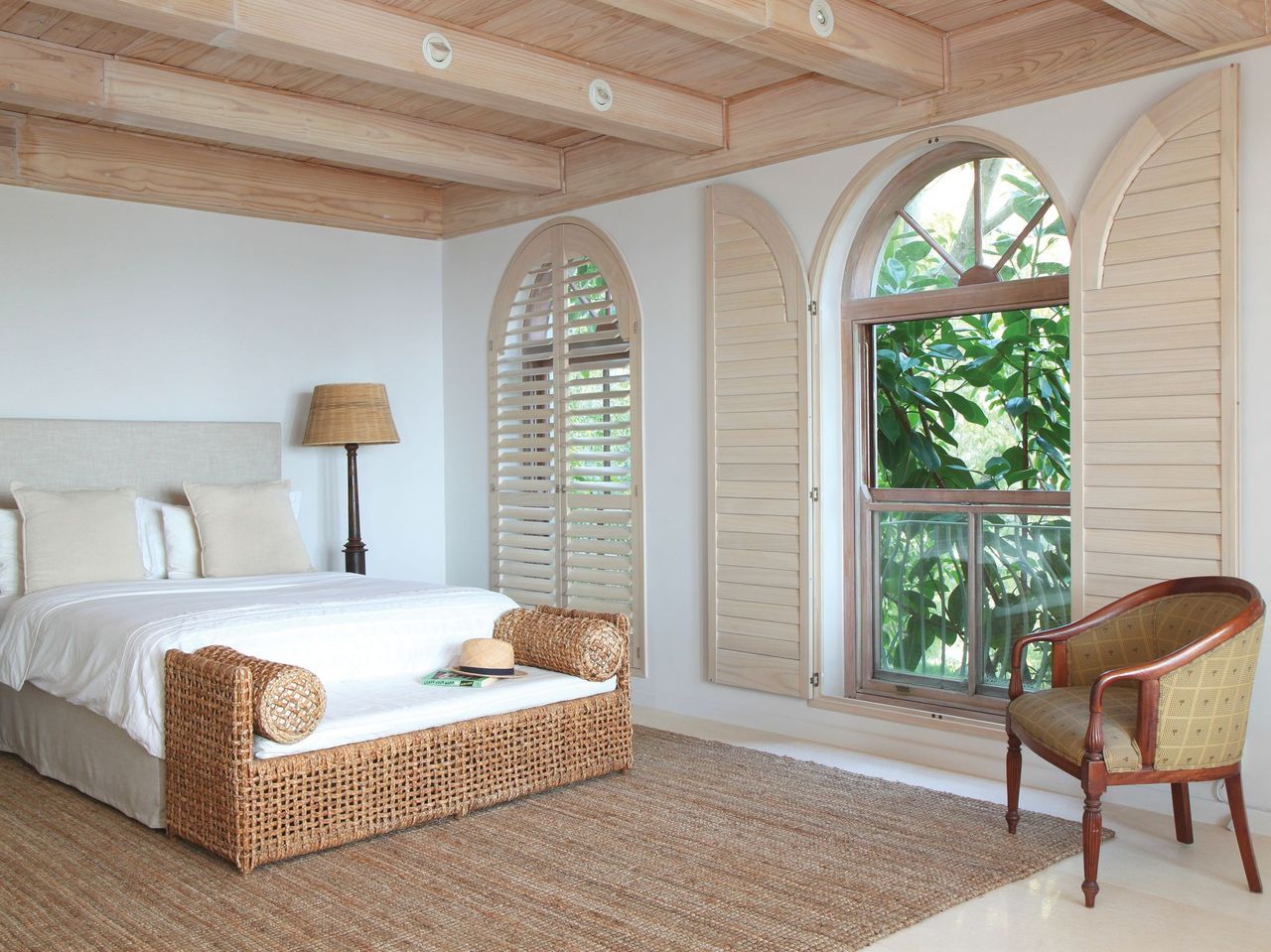 A bedroom with a woven bench at the foot of the bed, arched windows with shutters, and a wooden ceiling.