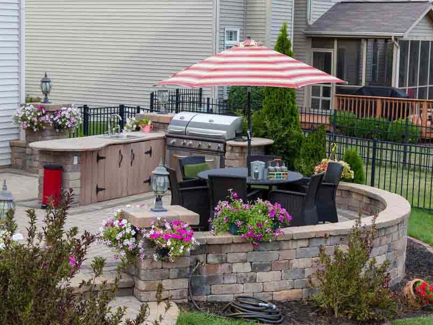 A patio with a stone outdoor kitchen, grill, and dining table under a red-and-white striped umbrella with flower planters.