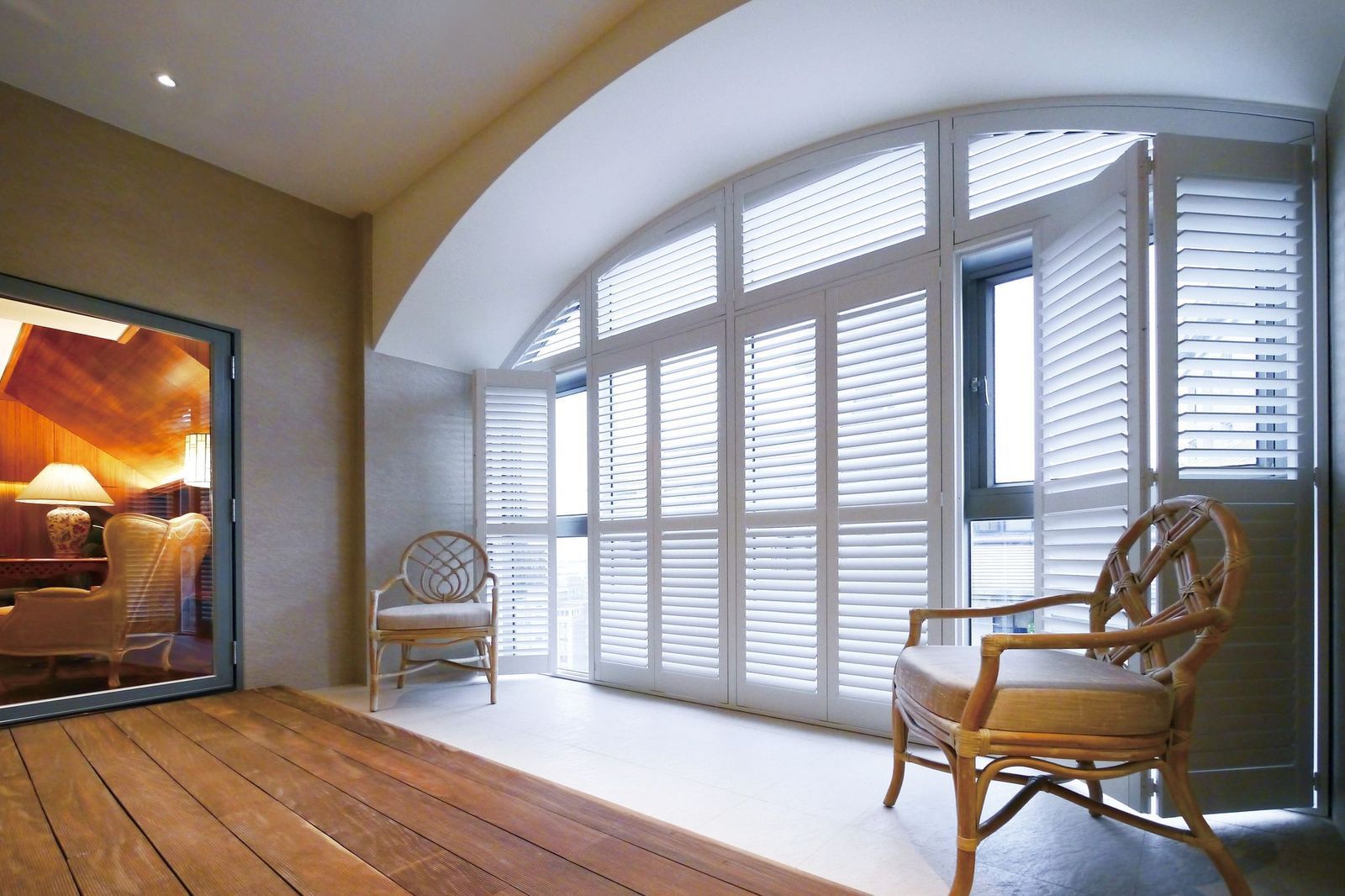 A sunroom featuring arched shutter windows, two wicker chairs, and a wooden floor, with a doorway leading into a room.