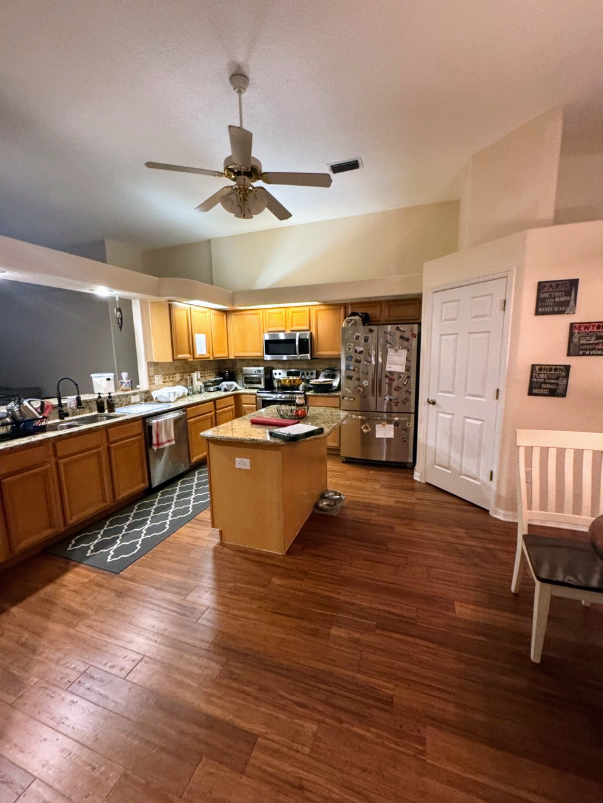 A modern kitchen featuring wooden cabinets, a central island, stainless steel appliances, and a ceiling fan.