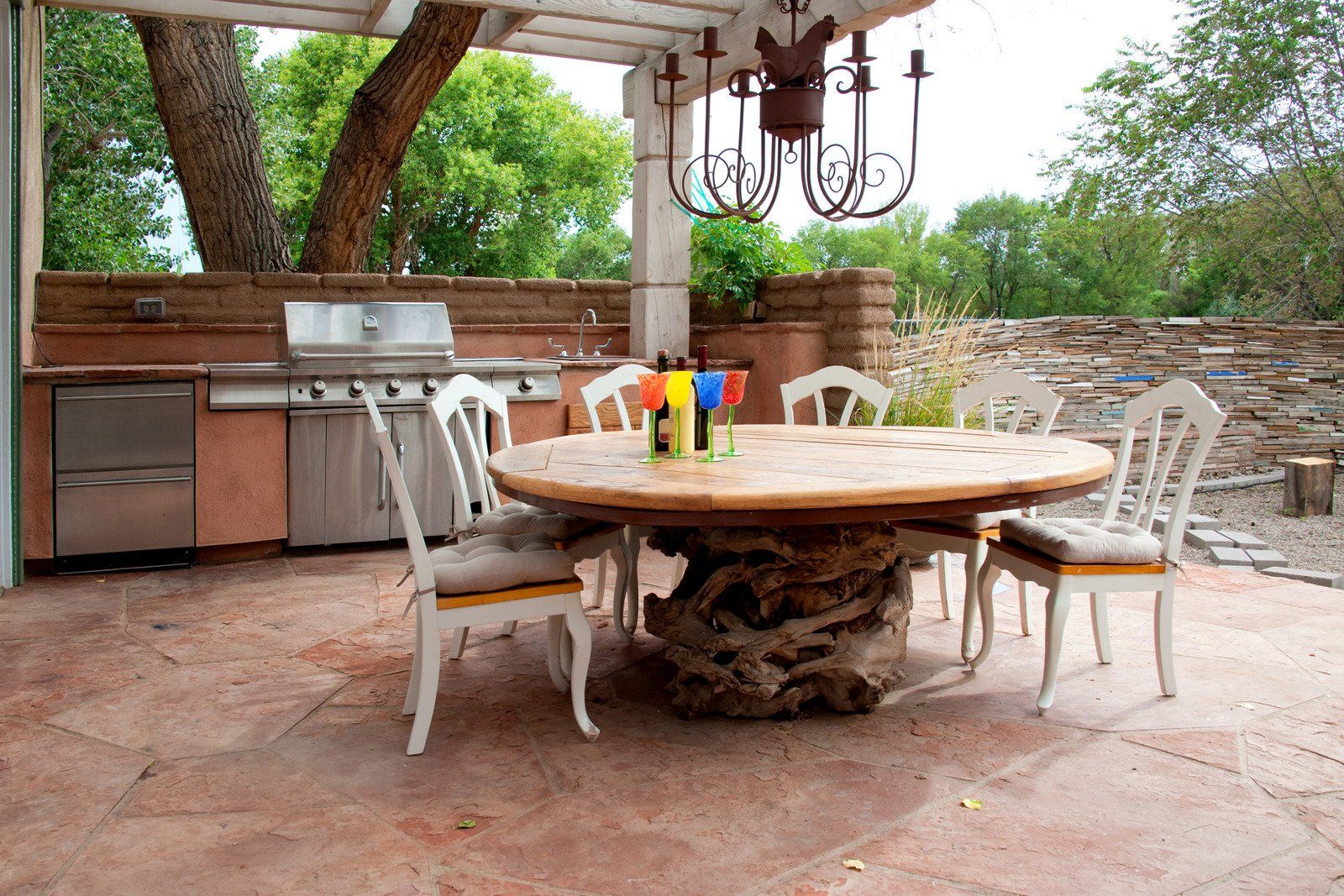 An outdoor patio with a circular table featuring a driftwood base, surrounded by white chairs, beside an outdoor kitchen.