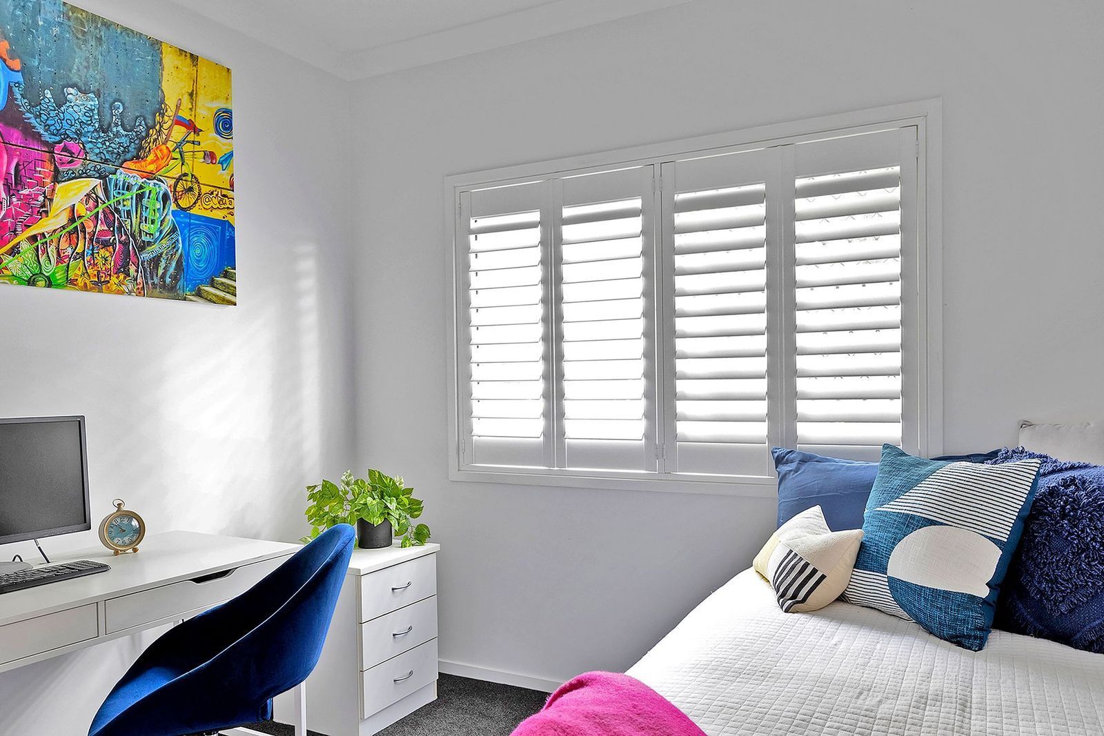A bedroom workspace with a desk, a blue chair, a potted plant, a bed with blue patterned pillows, and a vibrant painting.