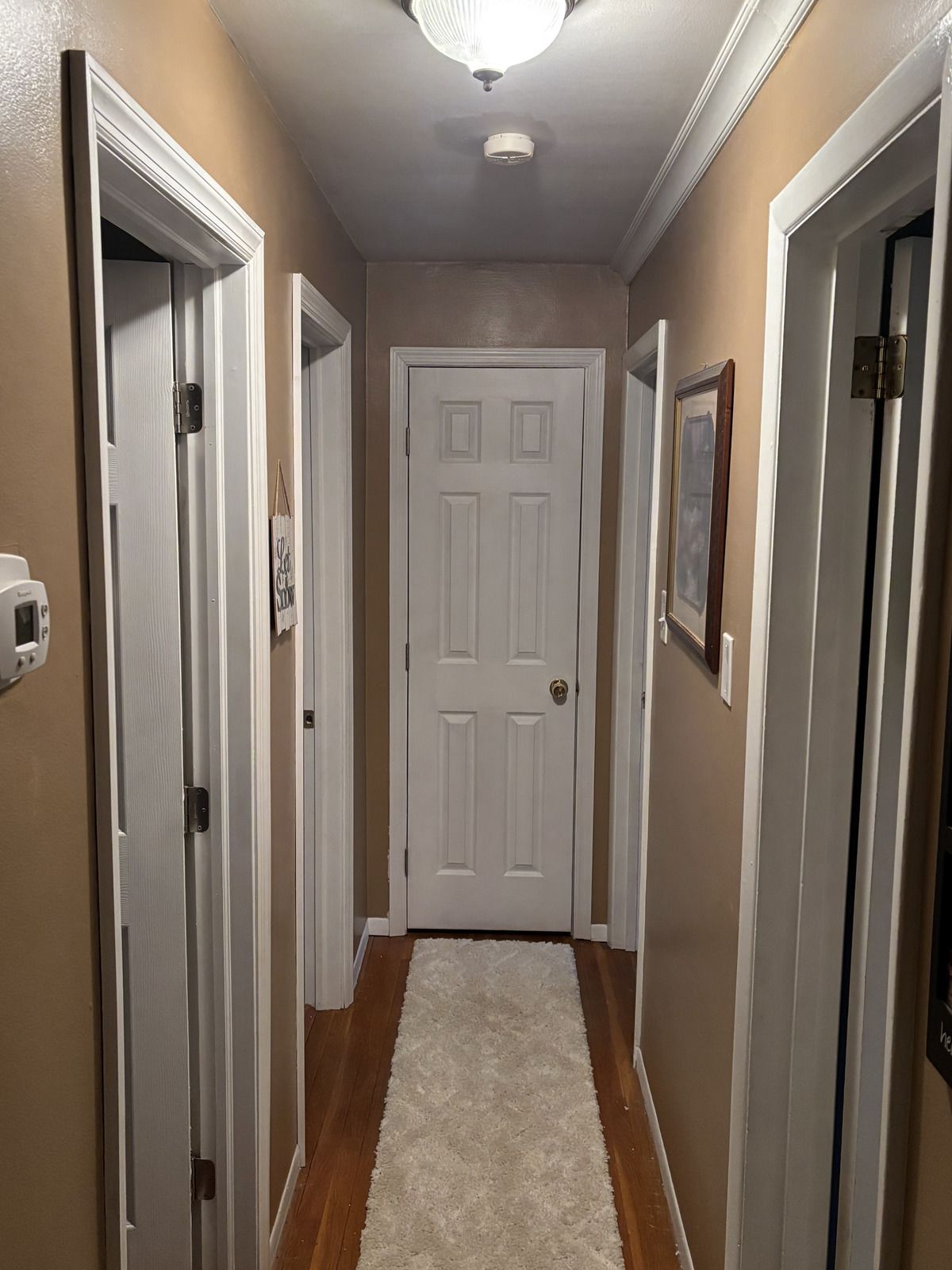 A narrow hallway with tan walls, white door frames, and a light-colored runner rug leading to a white door at the end.