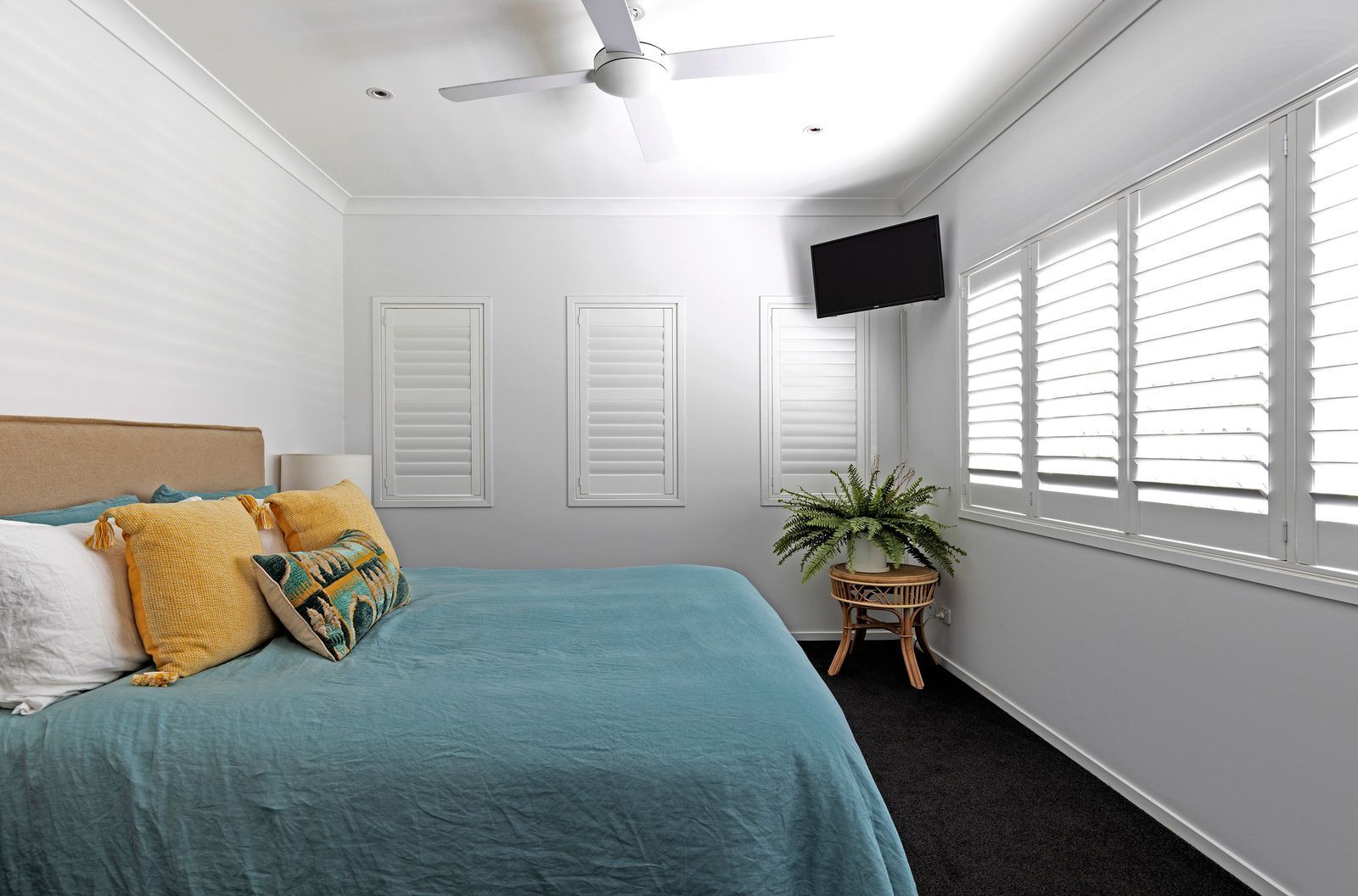 A bedroom with white walls, dark carpet, a teal bedspread, mustard and patterned pillows, and white shuttered windows.