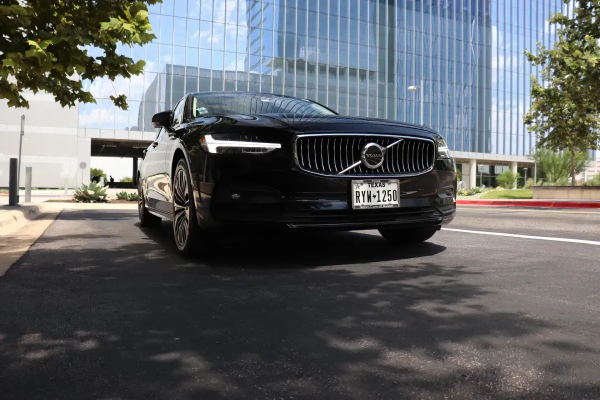 Black Volvo sedan driving on asphalt, in front of modern glass building. Texas license plate visible.