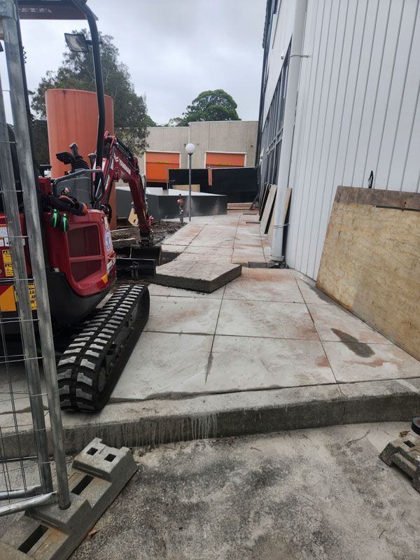 A small red excavator is sitting on a sidewalk next to a building.