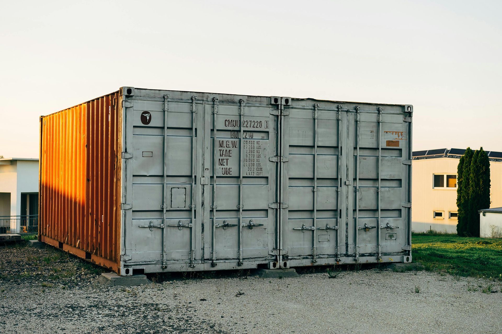 Grijze zeecontainer met oranje zijpanelen op een grindterrein bij zonsondergang.