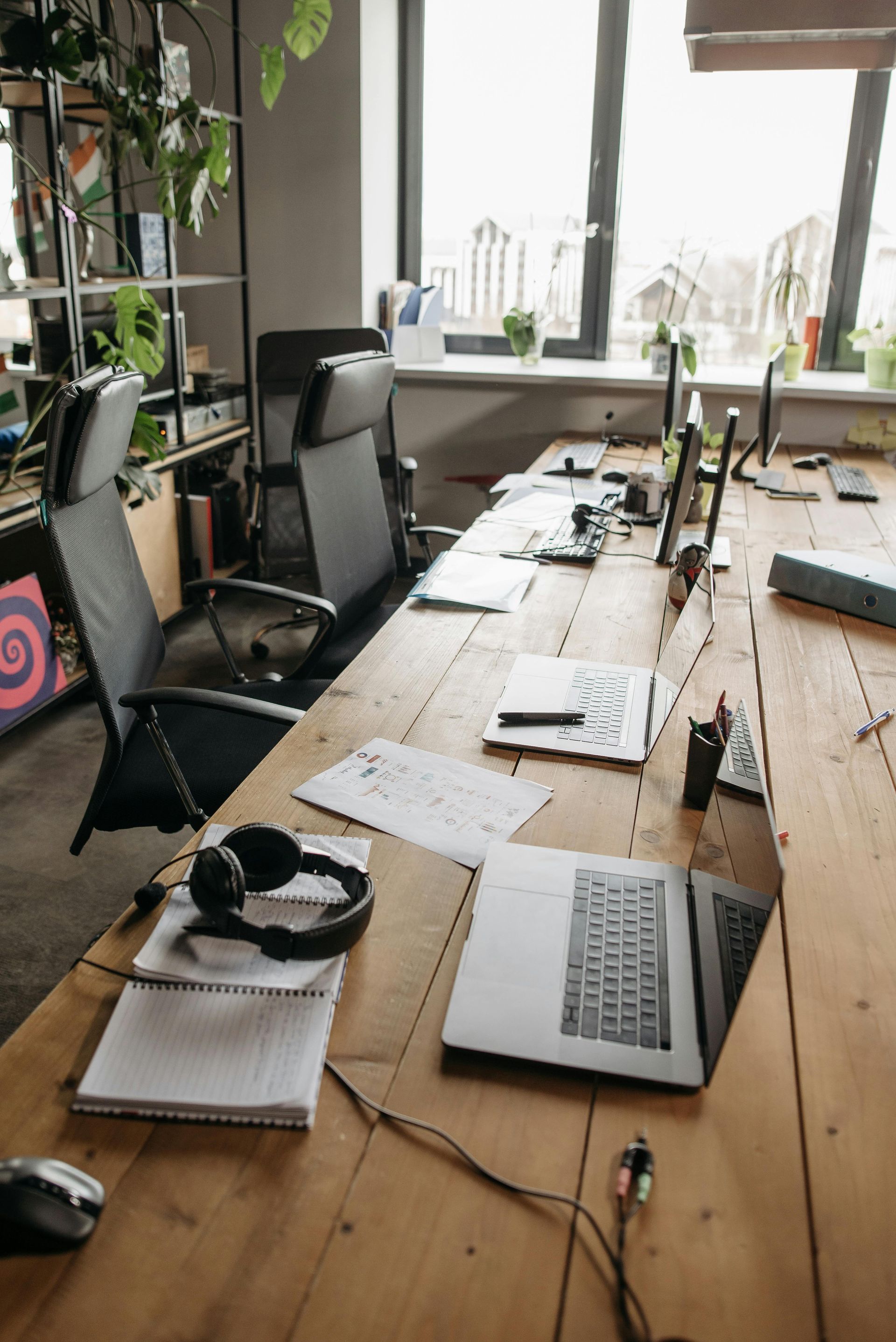 Een rommelig bureau in een thuiskantoor met laptop, koptelefoon, planten en papieren bij een licht raam.