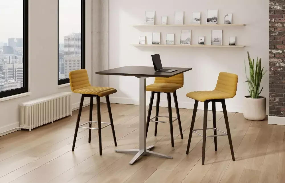 A modern office setting with a square table, yellow stools, and a laptop.