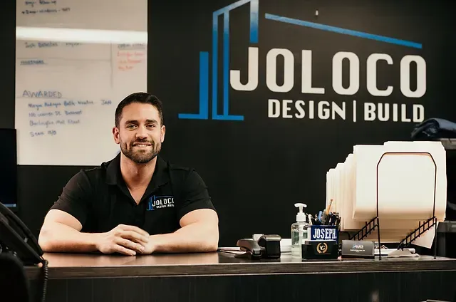A man is sitting at a desk in front of a wall that says joloco design build