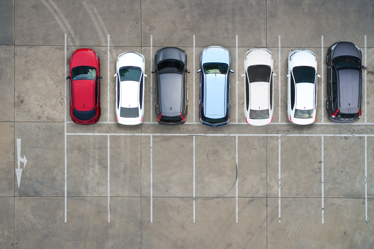 Seven parked cars in a row on a mostly empty parking lot, viewed from above.