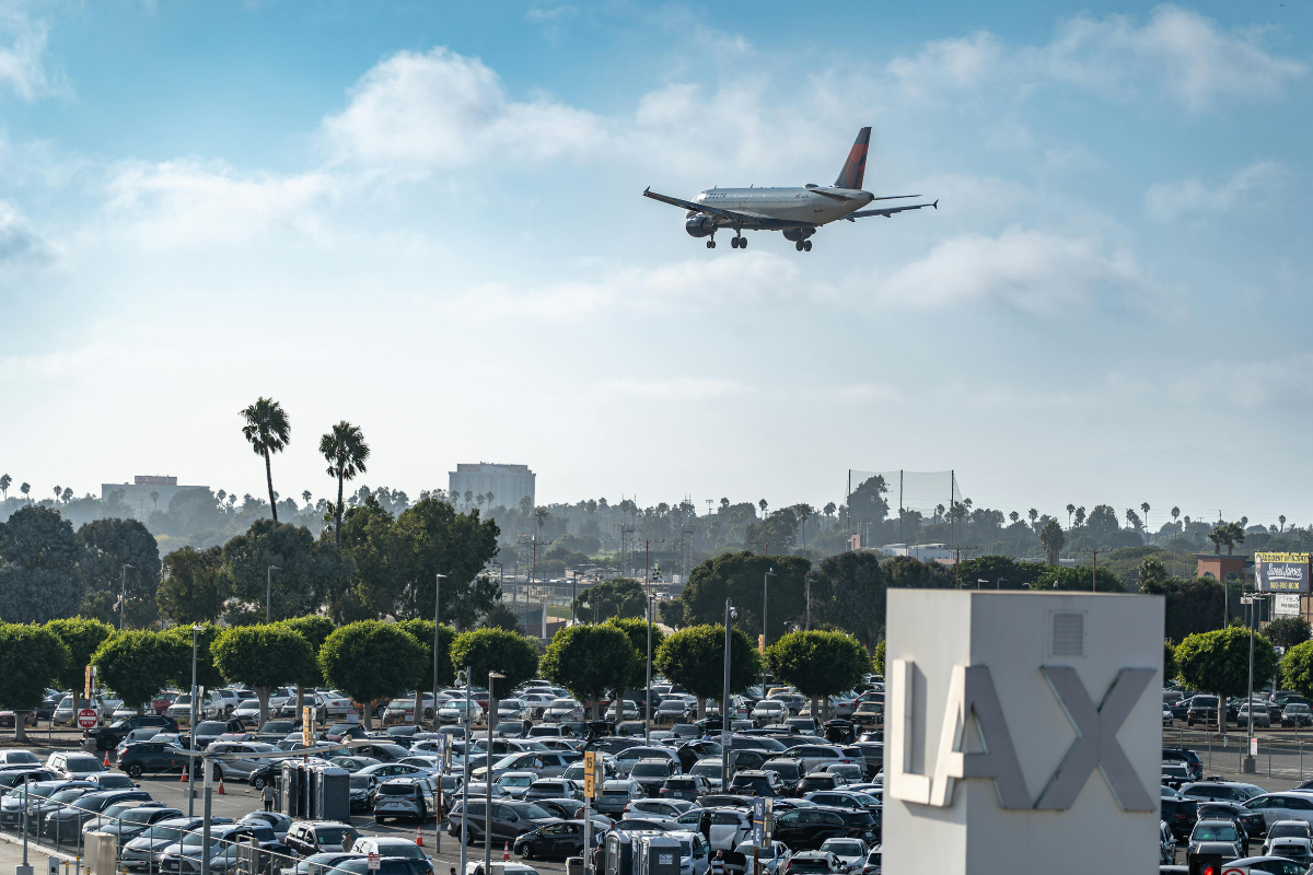 Airplane approaching LAX over a busy parking lot and city skyline under a blue sky