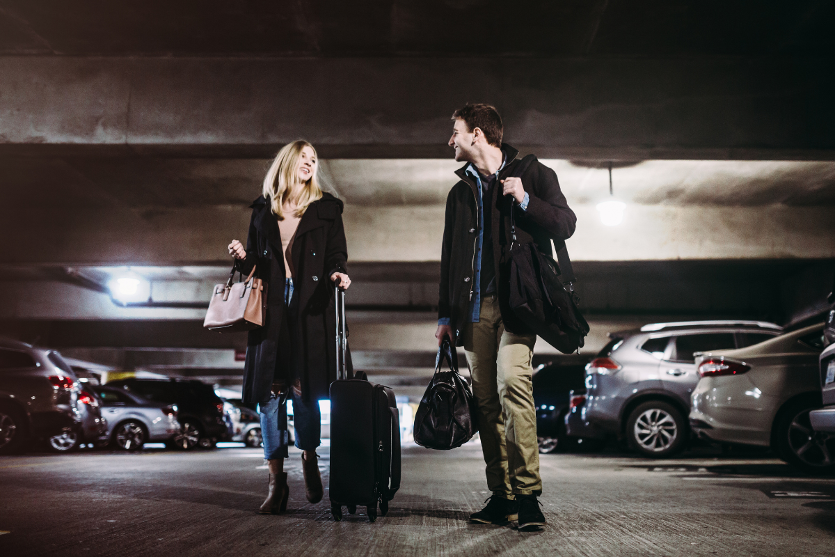 Two travelers with luggage talking in a dim parking garage beside parked cars