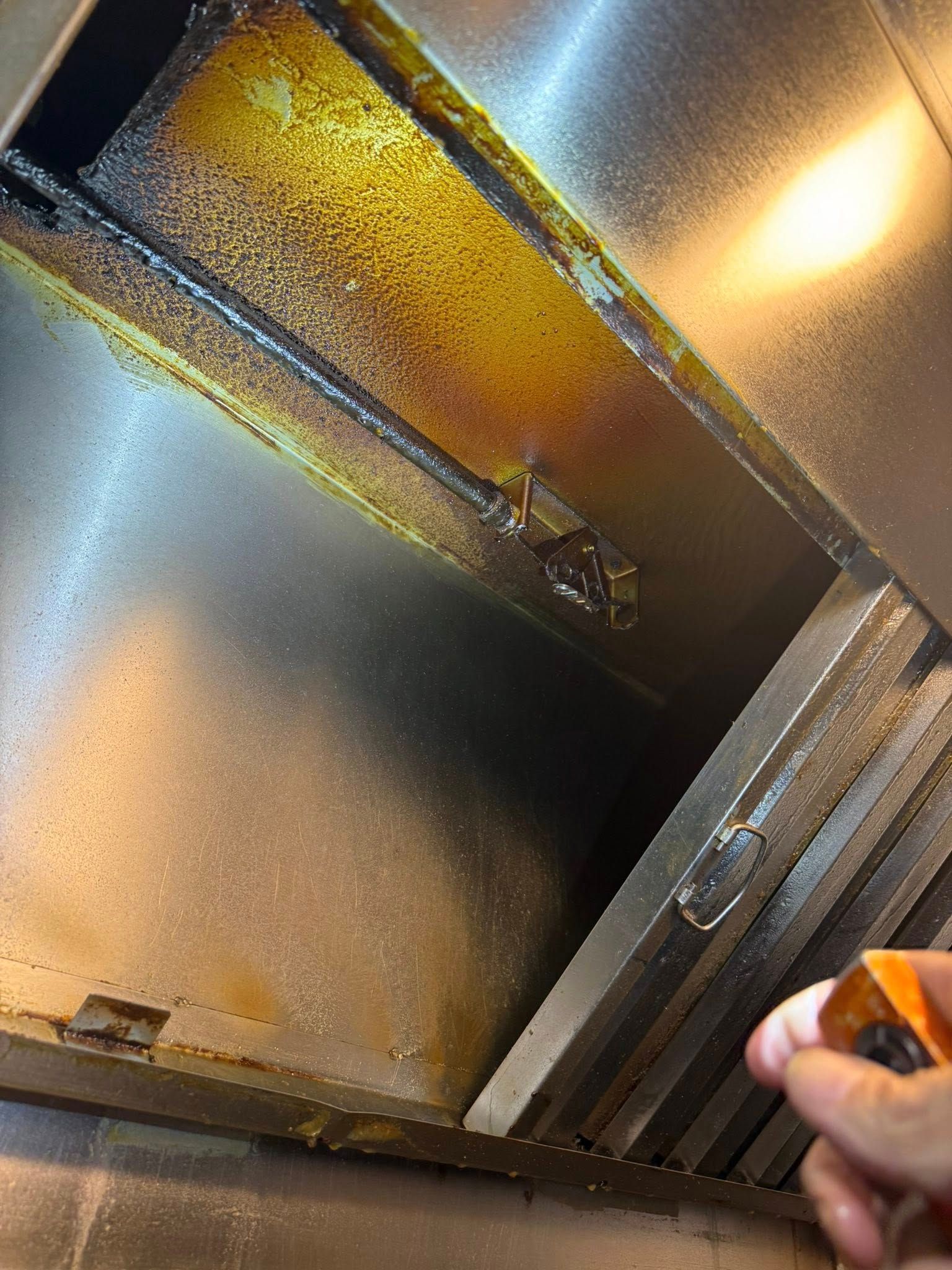 Hand holding a spray nozzle aimed at a metallic ventilation hood with grease buildup.