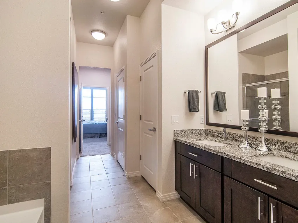 Bathroom vanity with double sinks, granite countertops, and dark wood cabinets, leading to a bedroom.