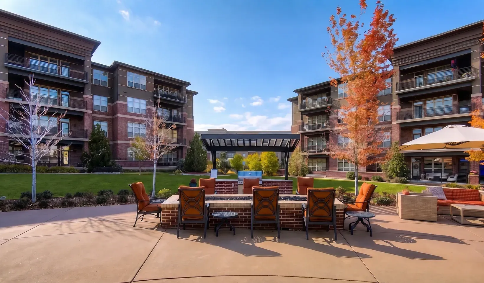 Outdoor courtyard with patio seating between apartment buildings and autumn trees, under a blue sky.