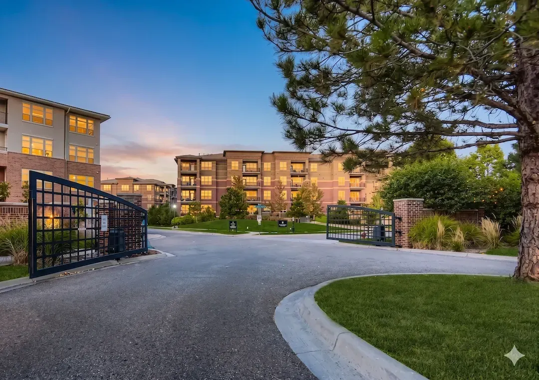 Gated apartment complex entrance at dusk with lit buildings, trees, and a paved driveway.