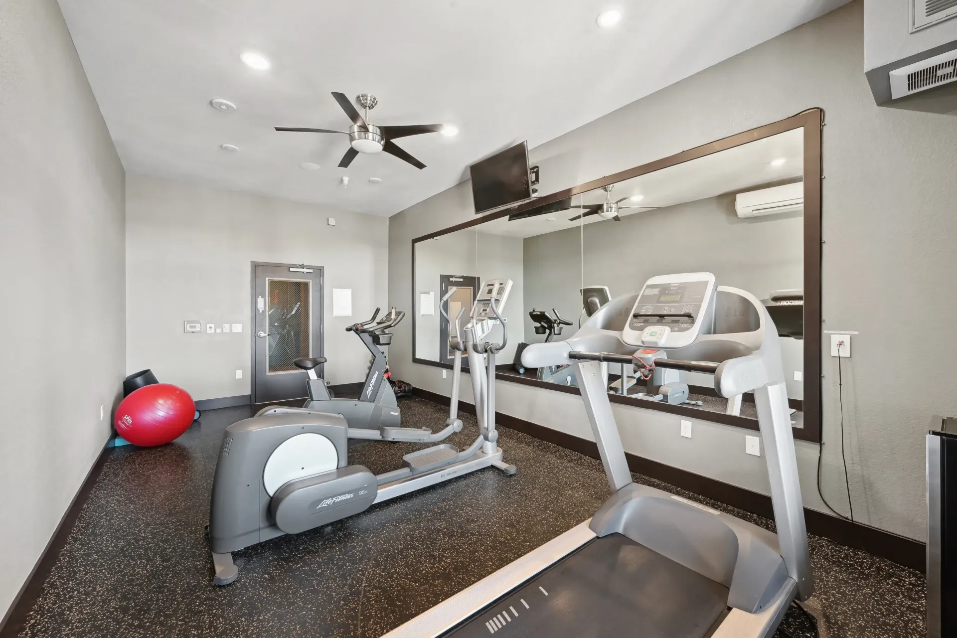 Home gym with treadmill, elliptical, exercise bike, wall mirror, and red stability ball.