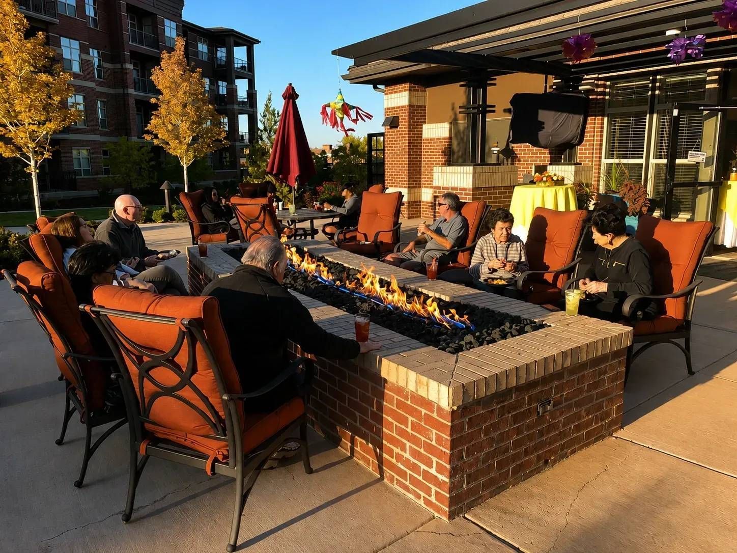 People relaxing around a brick fire pit on a sunny apartment patio with autumn trees nearby.