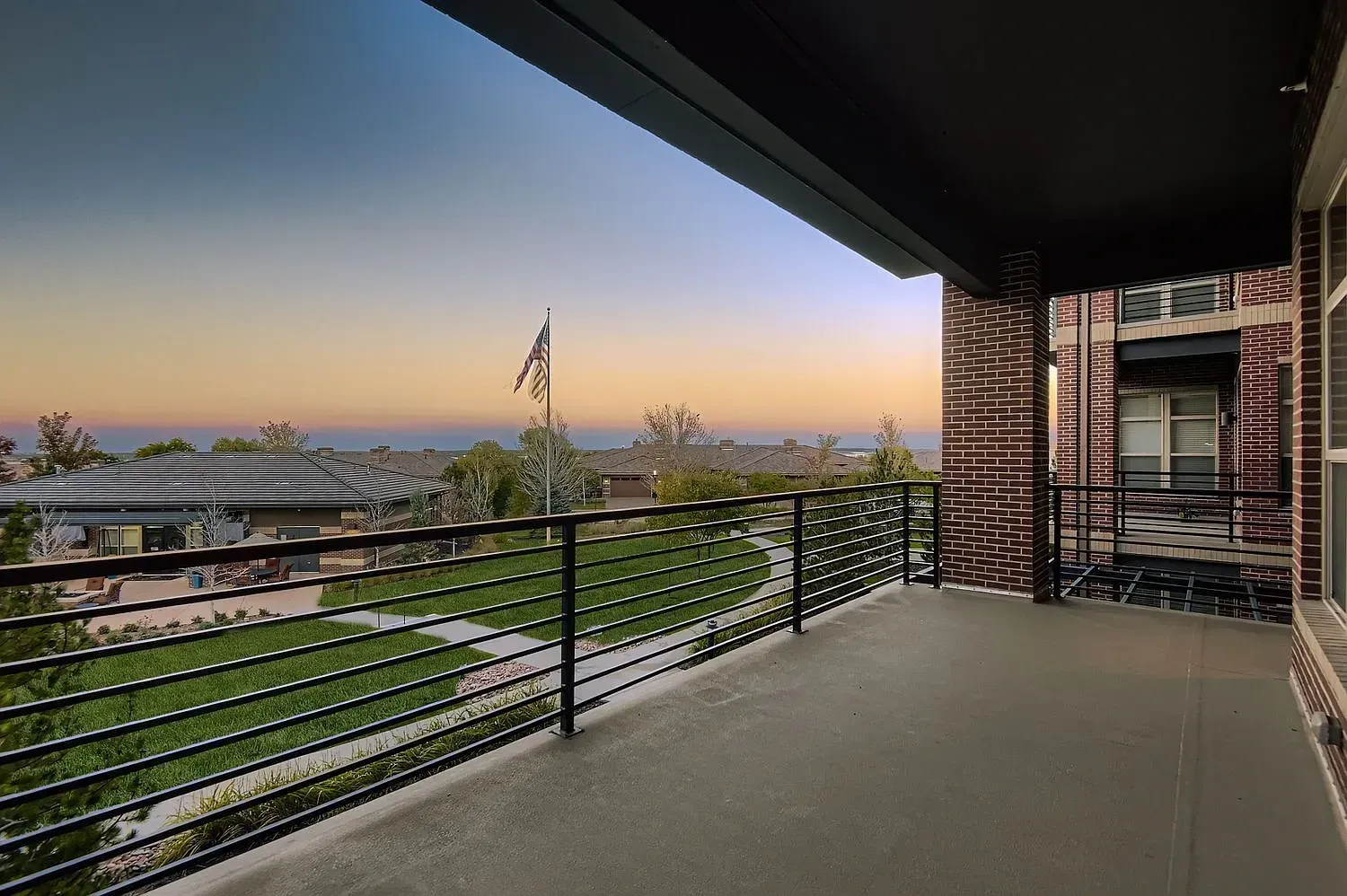 Balcony with black railing overlooking a sunset city view, framed by brick and wood exterior walls.