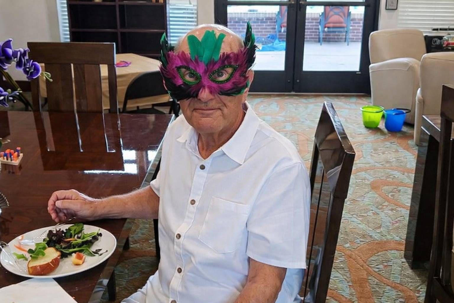 Person in a colorful mask sits at a dining table with a plate of food in a bright room.