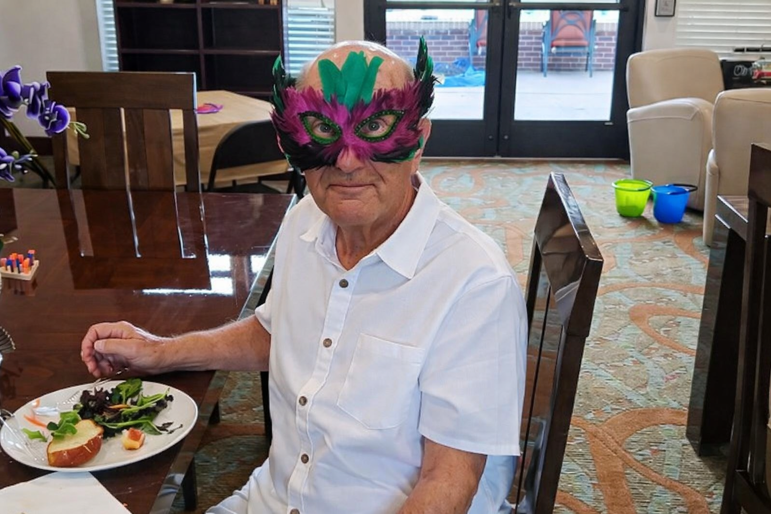 Person in a colorful mask sits at a dining table with a plate of food in a bright room.