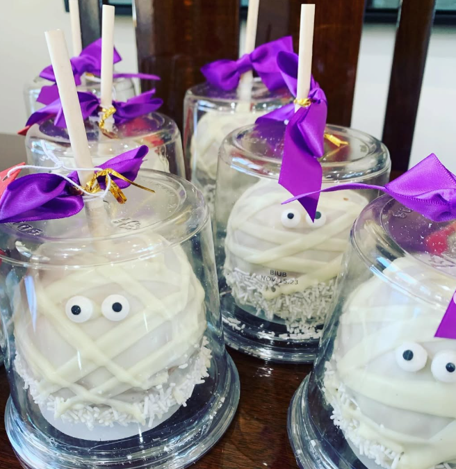 White mummy-themed treats in clear cups with purple bows and googly eyes on a table.