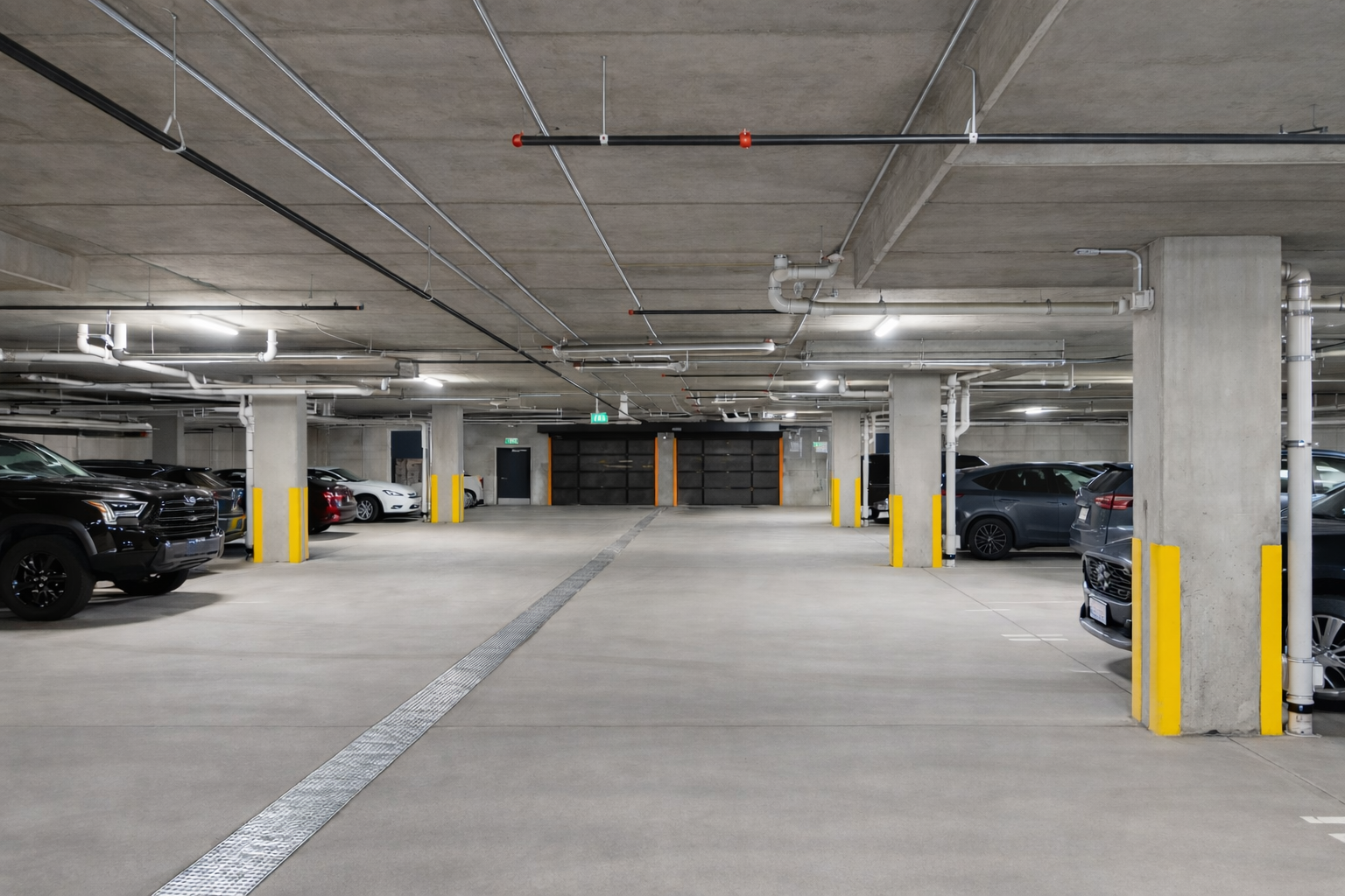Empty underground parking garage with concrete pillars, yellow bollards, and parked cars.