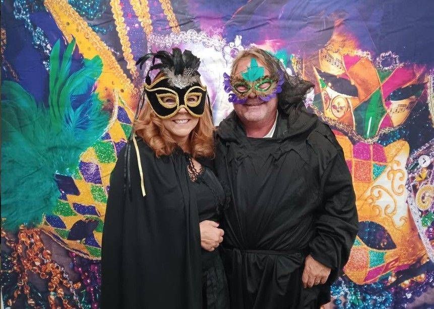 Two masked residents in black cloaks pose before a bright colorful mural for a holiday party.