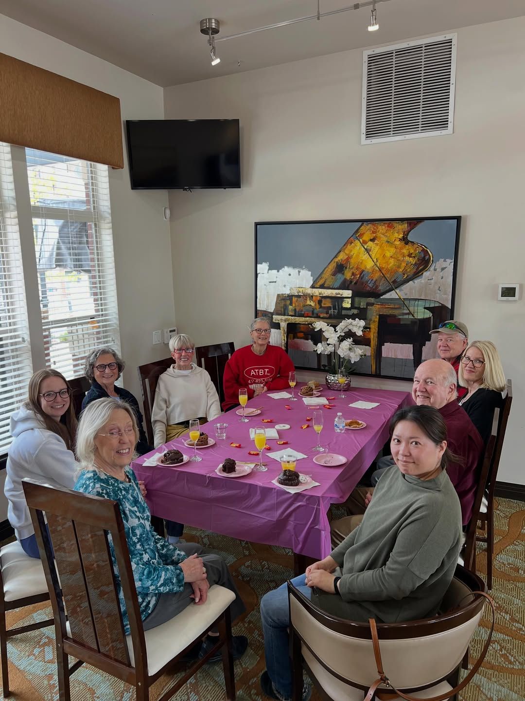 Group gathered around a purple-covered table in a bright dining room, smiling and seated for a meal.
