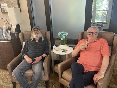 Two people sit in armchairs beside a small table in a lounge, one holding a drink.