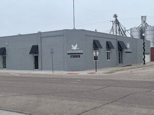 Gray industrial building with a sign, small awnings, and silos in the background along a street corner.