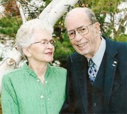 Two smiling adults standing outdoors in front of a tree, one in a green jacket and one in a dark suit.