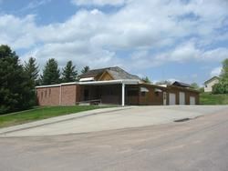 Brick community building with a covered entrance and garage bays beside a sloped driveway under a cloudy sky