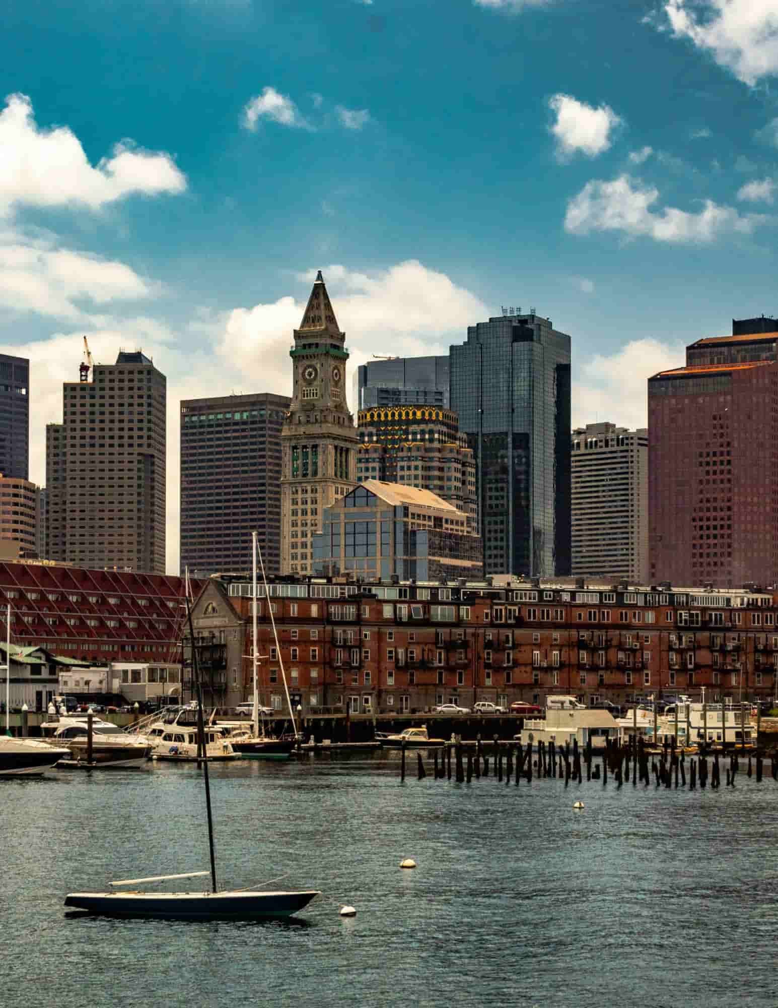 Boston skyline viewed from the waterfront, featuring historic Long Wharf, sailboats in the harbor, and the Custom House Tower rising above downtown.