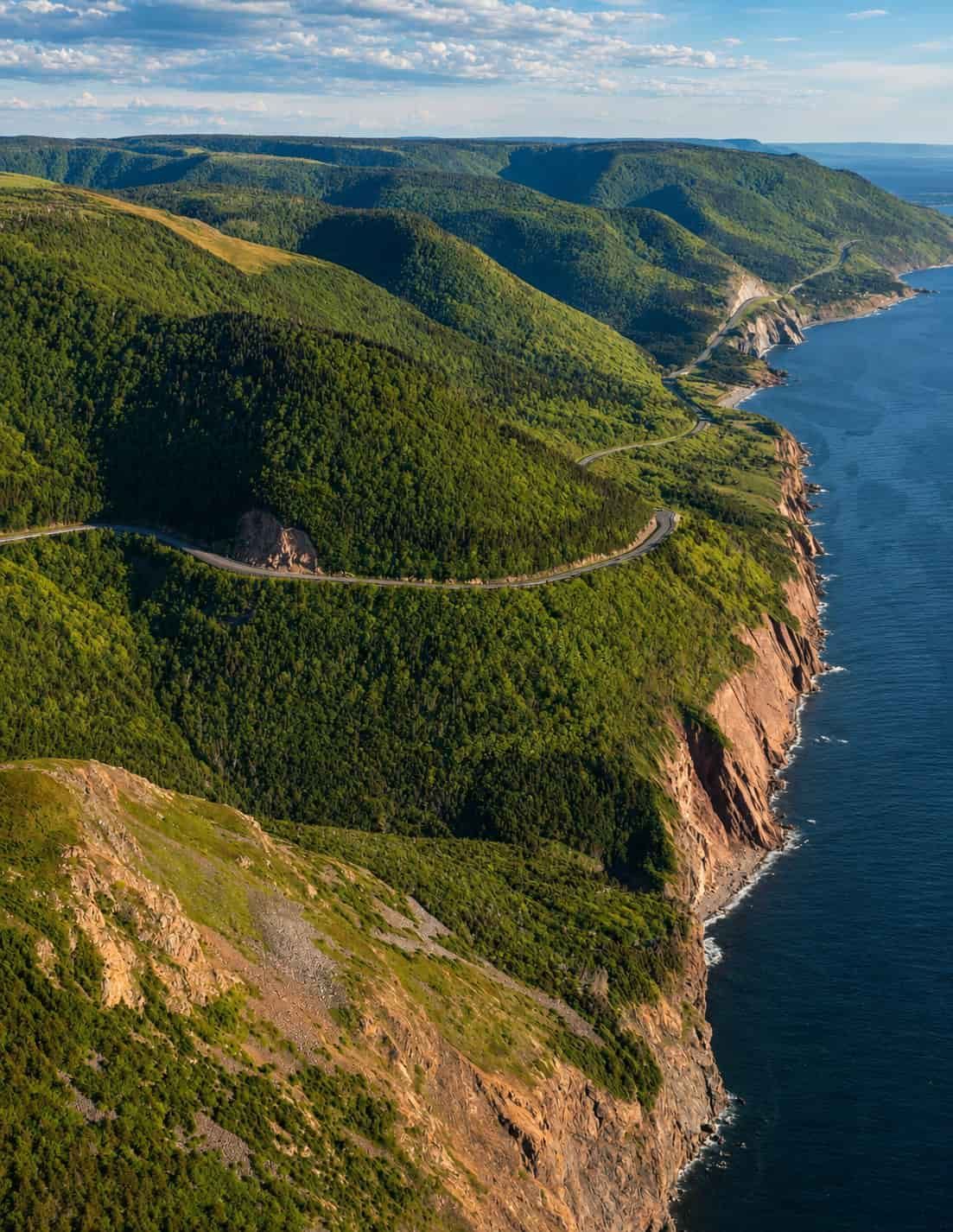 Aerial view of the Cabot Trail winding through Cape Breton’s forested highlands beside the Atlantic Ocean.