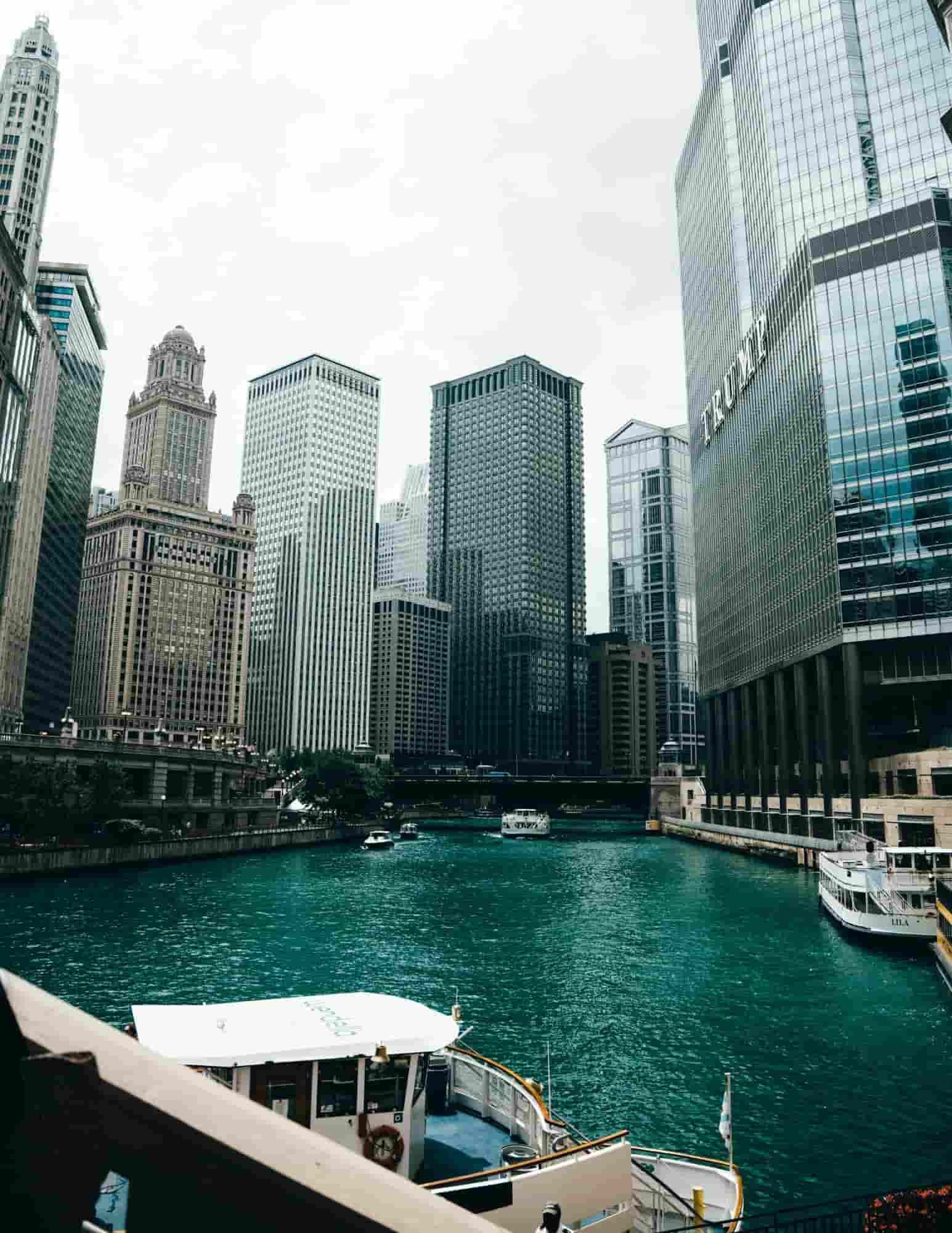 View of the Chicago River surrounded by downtown skyscrapers, with tour boats on the water and the city's iconic skyline under an overcast sky.