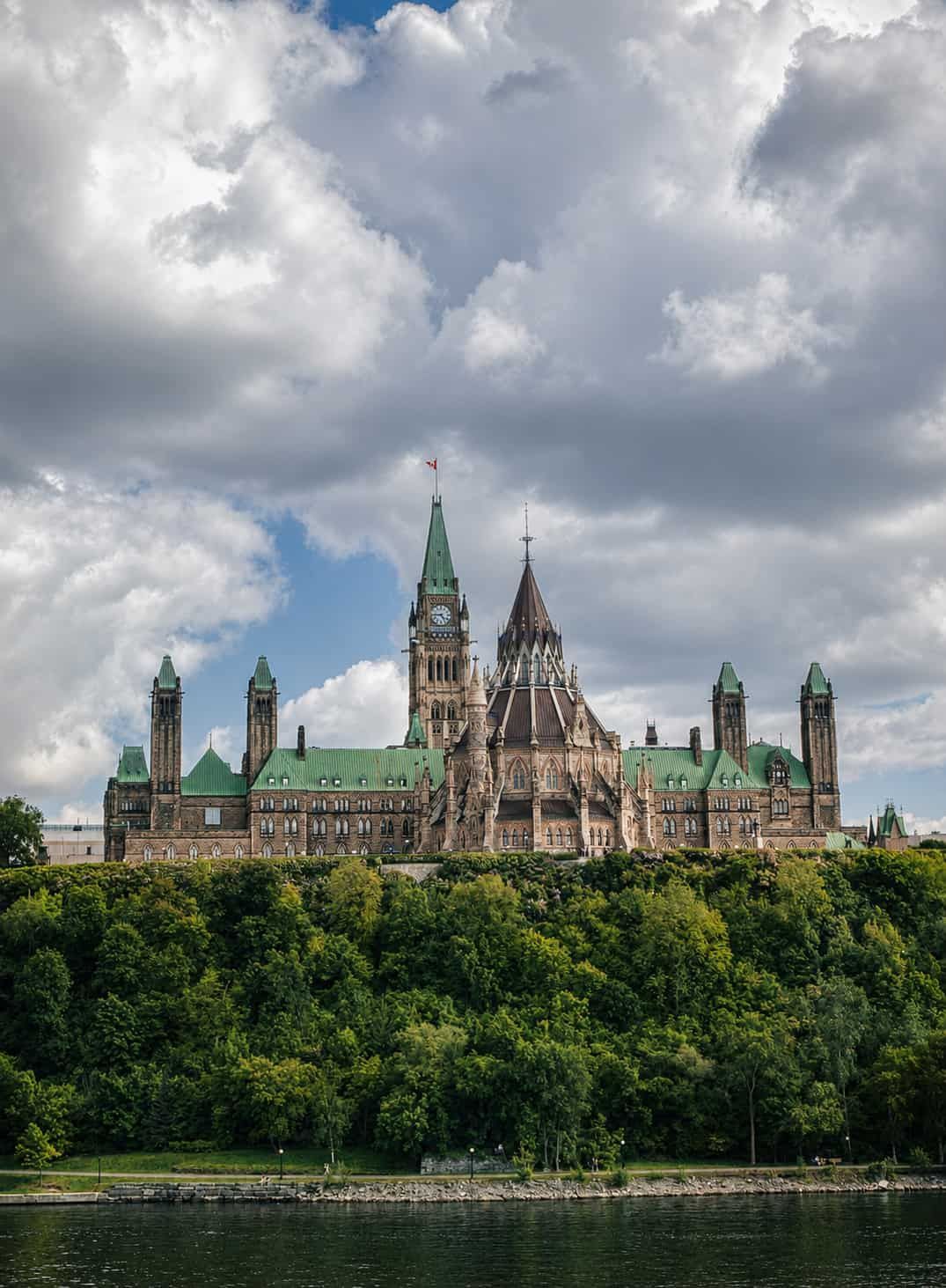 Parliament Hill in Ottawa overlooking the river beneath dramatic clouds, surrounded by lush green trees.