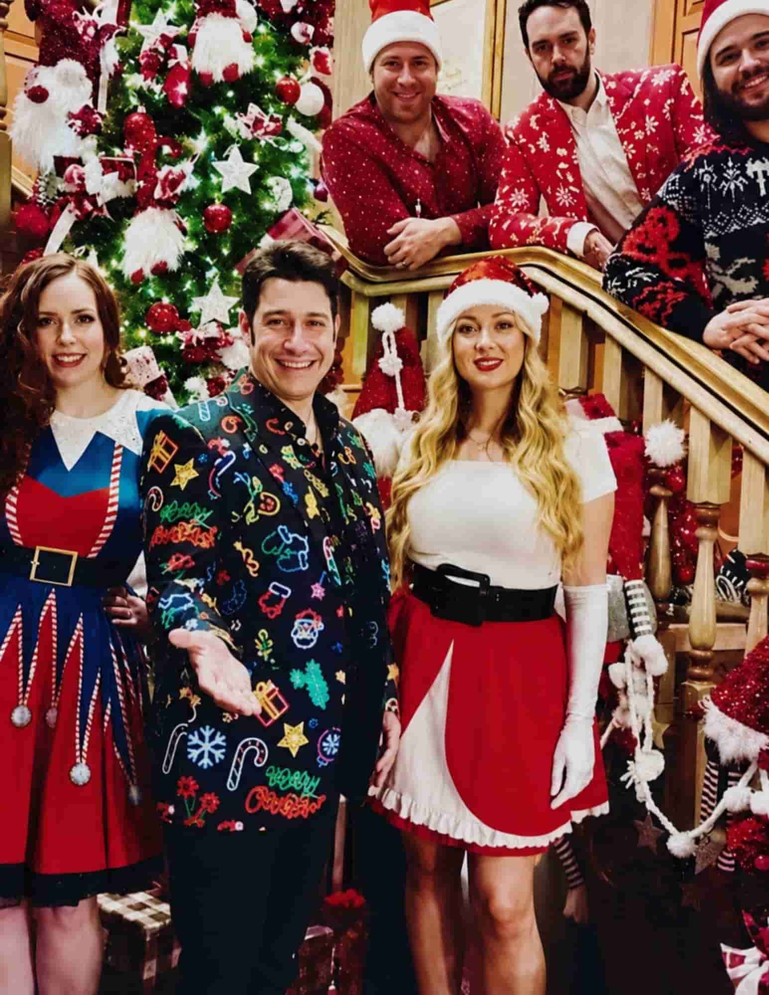 A group of six adults dressed in festive Christmas outfits pose on a decorated staircase beside a brightly lit Christmas tree.