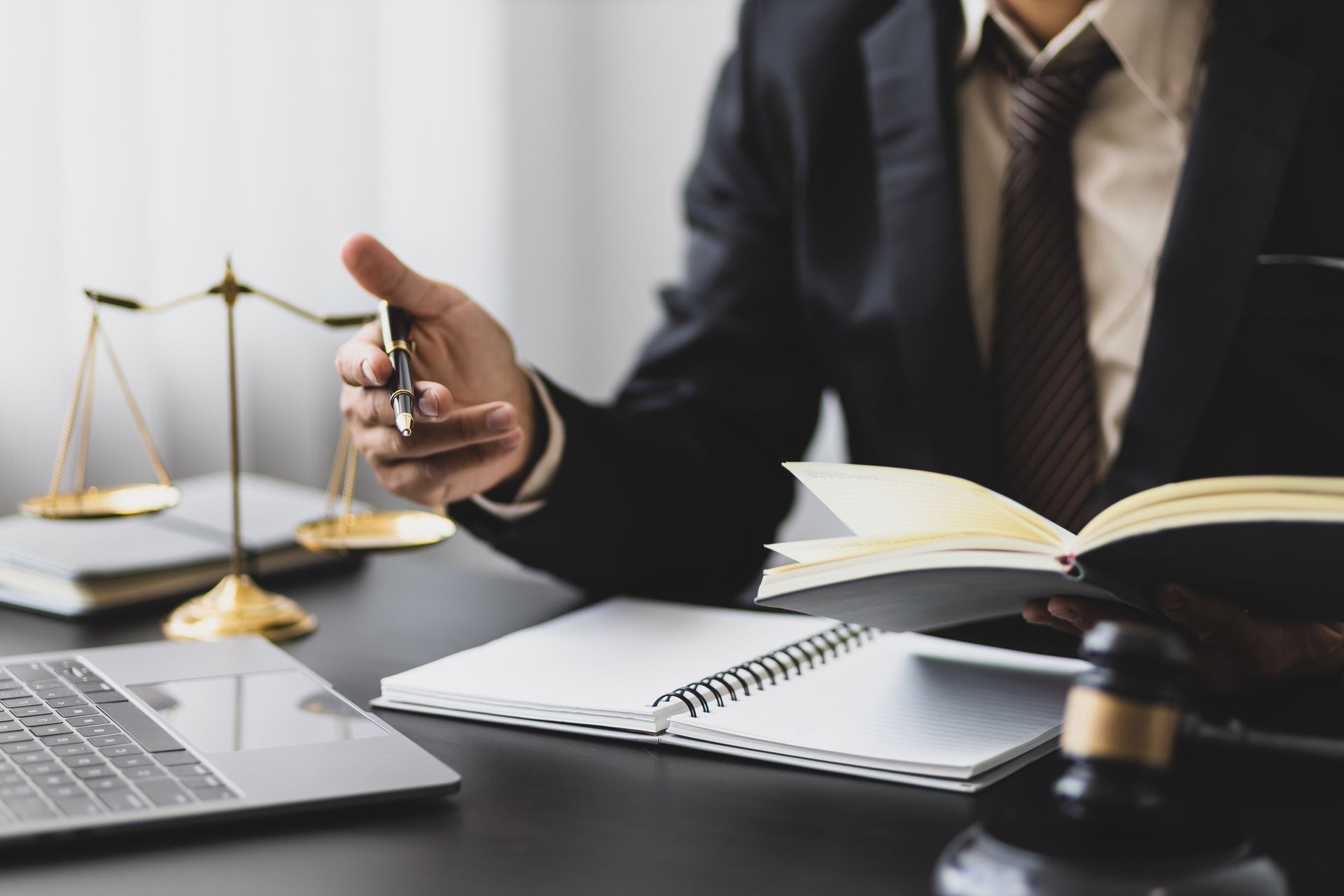 man in suit sitting at desk. Desk is covered with journals, laptop, and scales of justice.