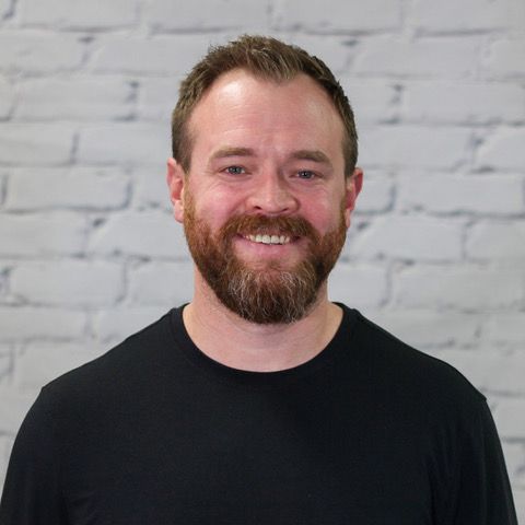 A man with a beard is wearing a grey shirt and smiling.
