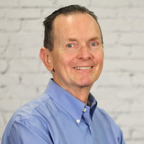 A man in a blue shirt is smiling in front of a white brick wall
