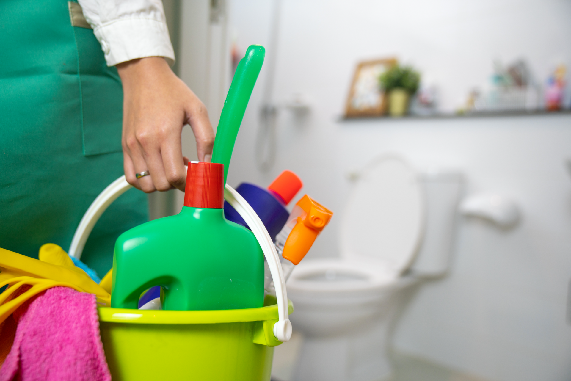 Person holding cleaning tools in a green bucket beside a toilet and bathroom supplies