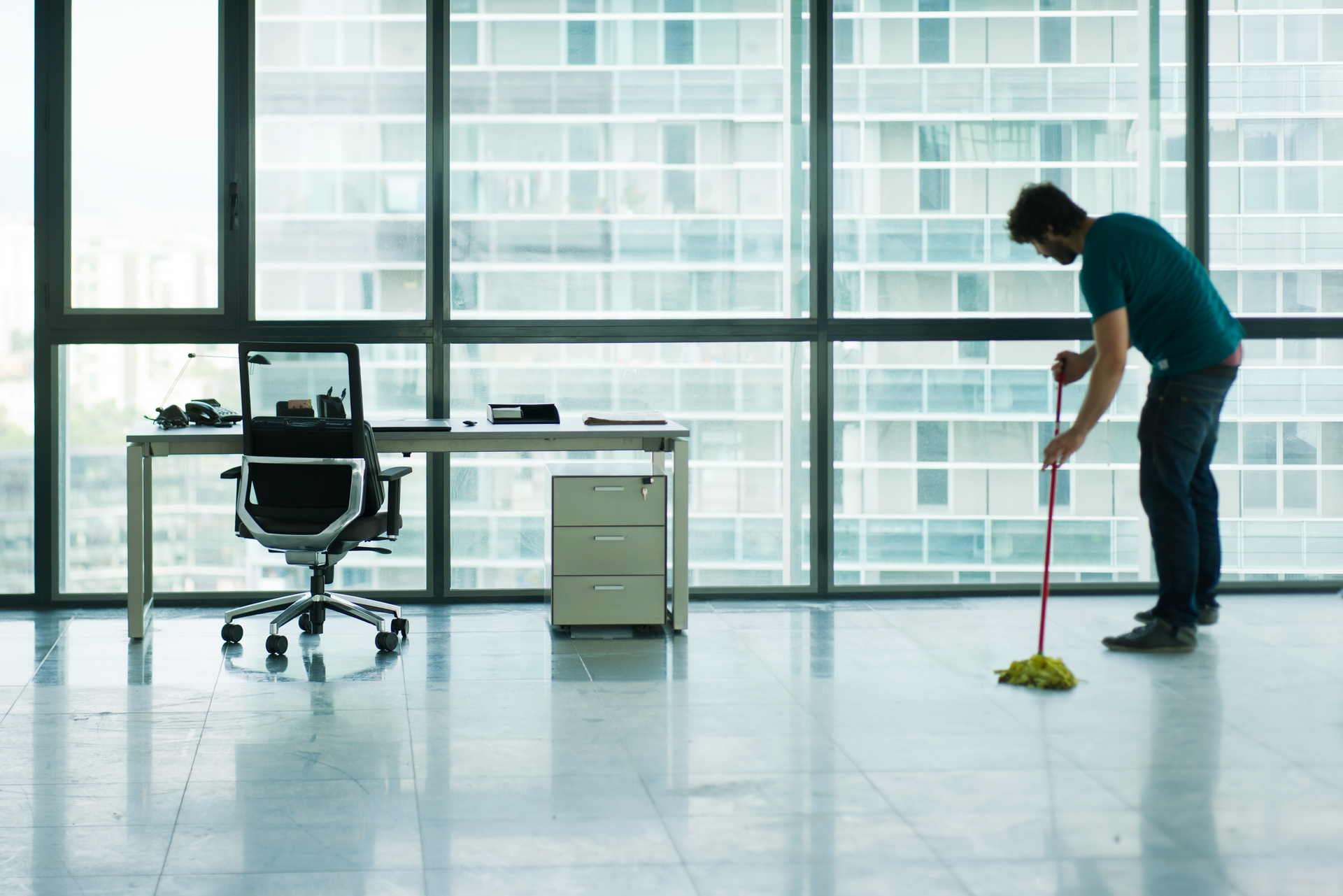Person mopping a glossy office floor beside a desk and chair by large windows